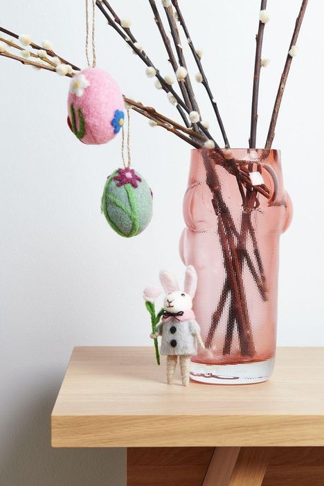 Zwei Filz-Ostereier an Zweigen in einer rosa Vase, daneben steht ein Filzkaninchen mit Schal und Blumen.