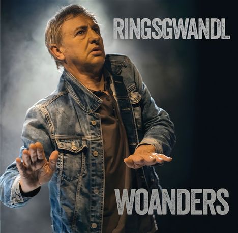 Text: "RINGSGWANDL" und "WOANDERS". Eine Person in Jeansjacke hebt die Hände, ein nachdenklicher Ausdruck.