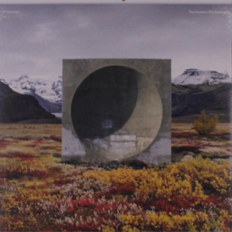 Text: "The Human...". Ein quadratischer Betonblock mit Kreisöffnung steht in einer bunten Landschaft mit Bergen im Hintergrund.