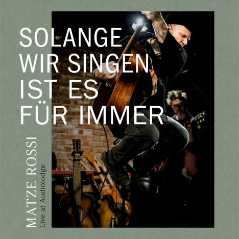 "Solange wir singen ist es für immer. Matze Rossi Live at Audiolodge." Ein Musiker springt mit Gitarre vor Backsteinwand.