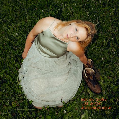 Text: "Karl die Große IST NICHT AUFGESCHOBEN." Eine Frau im grünen Kleid sitzt im Gras neben Wanderstiefeln.