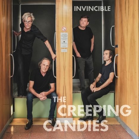 Text: "INVINCIBLE" oben, "THE CREEPING CANDIES" unten. Vier Personen in schwarzen Outfits stehen in einem holzgetäfelten Aufzug.
