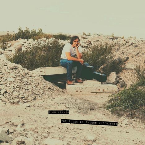 "Slow Leaves" und "The Ruins of Things Unfinished." Ein Mann sitzt nachdenklich auf Trümmern, umgeben von Schutt und Grün.