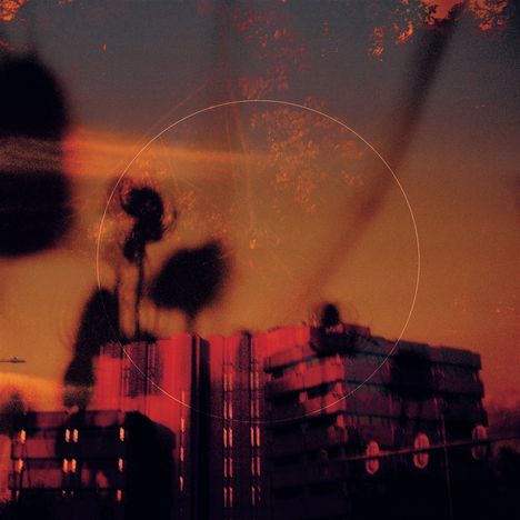 Eine verträumte Szene zeigt einen rötlich beleuchteten Gebäudekomplex und verschwommene Silhouetten von Pflanzen im Vordergrund.