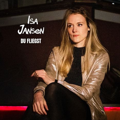 Text: "Isa Jansen - Du fliegst". Eine Frau in goldener Jacke sitzt, Blick zur Seite, in dunklem Umfeld mit rotem Licht.