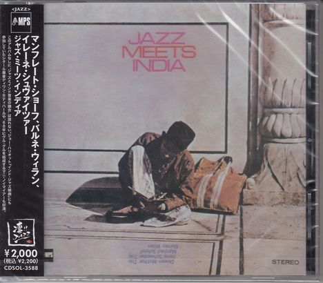 "JAZZ MEETS INDIA" in rosa Schrift. Ein Mann sitzt neben einem großen Kissen oder Sack, in ruhiger Umgebung.