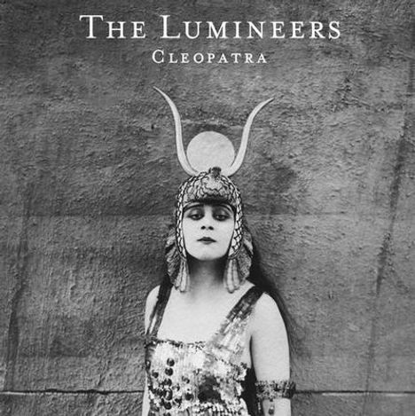 "The Lumineers", "Cleopatra". Schwarzweißfoto einer Frau in antikem Kostüm mit Kopfschmuck vor einer schlichten Wand.