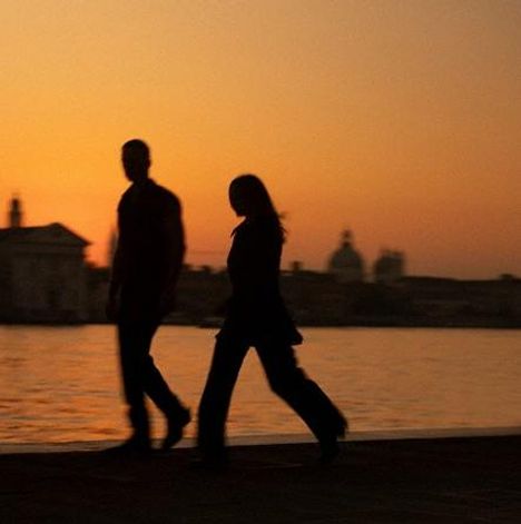 Zwei Personen gehen vor einem Sonnenuntergang, im Hintergrund eine Stadtsilhouette und ein Gewässer.