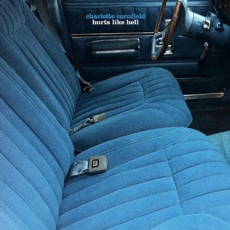 Text: "charlotte cornfield hurts like hell." Ein Auto mit blauen, gesteppten Sitzbezügen und Lenkrad aus Holz.