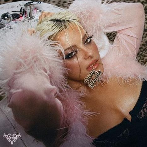 Text: "Naughty Blonde" auf einem Kettenanhänger. Frau mit blonden Haaren in rosa, flauschigem Outfit auf einem Bett.