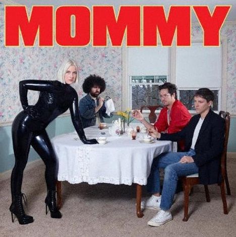 Text "MOMMY" in roter Schrift oben. Drei Männer sitzen am Tisch, eine Frau in schwarzem Outfit steht daneben.