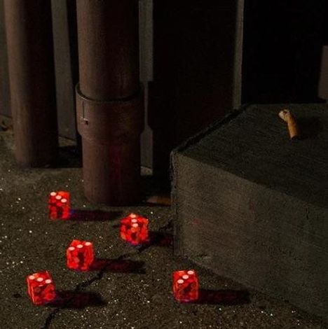Rote Würfel auf Asphalt, eine Zigarettenkippe liegt auf einer Kante. Hintergrund metallisch und dunkel.