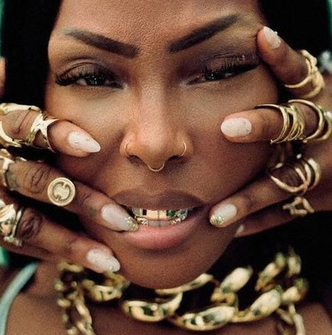 Kein Text vorhanden. Frau mit goldenen Accessoires, inklusive Nasenring und Zahnverzierung. Hände mit vielen Ringen umrahmen ihr Gesicht.