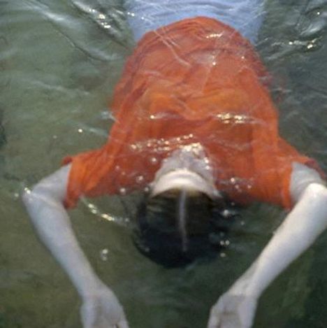 Eine Person in einem orangefarbenen Hemd treibt mit ausgestreckten Armen auf dem Rücken im Wasser.