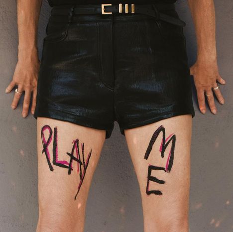 Schriftzug "PLAY ME" auf den Oberschenkeln einer Person in schwarzen Ledershorts.