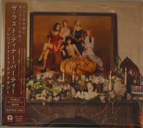 Japanischer Text auf der linken Seite. Im Hintergrund ein Gemälde mit fünf stilvoll gekleideten Personen. Vorne Kerzen und Blumen.