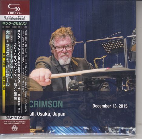 Texte: "King Crimson", "Osaka, Japan", "December 13, 2015". Eine Person mit Brille spielt Schlagzeug.