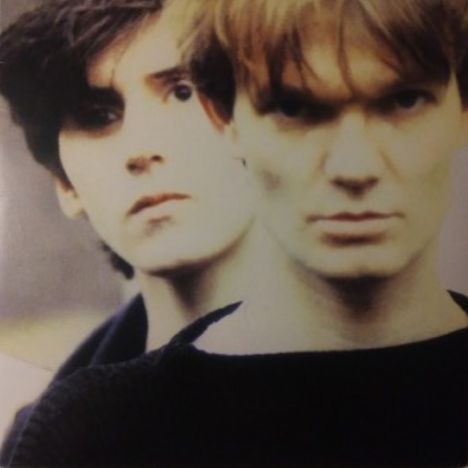 Zwei Personen blicken ernst in die Kamera. Der Vordergrund zeigt eine Person mit kürzeren Haaren, dahinter eine mit längeren.