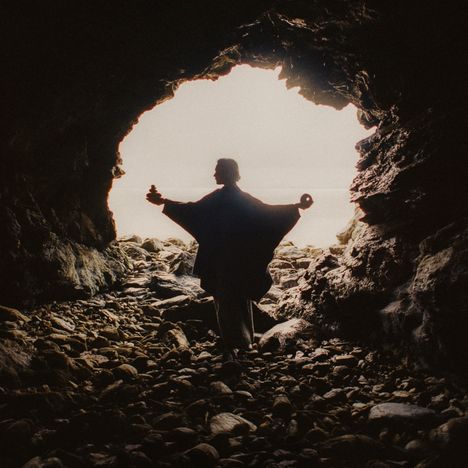 Eine Person steht in einer Höhle, siluettiert gegen das Licht, hält einen Stein-Stapel in der Hand, umgeben von Felsen.