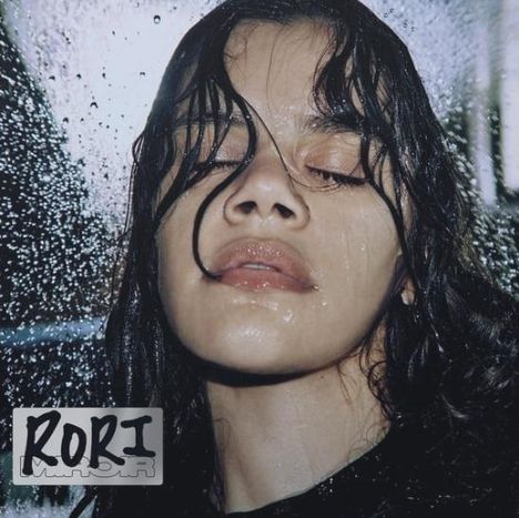 Text: "RORI". Eine Person mit nassen Haaren und geschlossenen Augen steht im Regen oder hinter einer regennassen Scheibe.