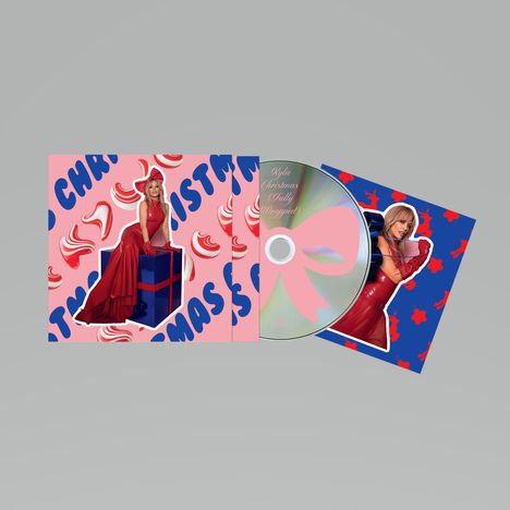 Text: "Kylie Christmas (Fully Unwrapped)". Rotes Kleid, Frau auf Geschenkbox, pink-blauer Hintergrund mit Schleifenelementen.