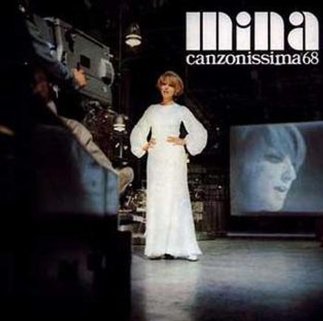 Text: "Mina canzonissima68". Eine Frau in einem langen weißen Kleid steht, Gesichtsausdruck ernst. Im Hintergrund Bildschirm.