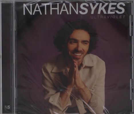 "NATHAN SYKES ULTRAVIOLET". Ein Mann lächelt und faltet die Hände, trägt ein helles Hemd vor dunklem Hintergrund.