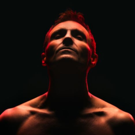 Dunkler Hintergrund, Oberkörper eines Mannes mit nach oben geneigtem Kopf, rot beleuchtet.