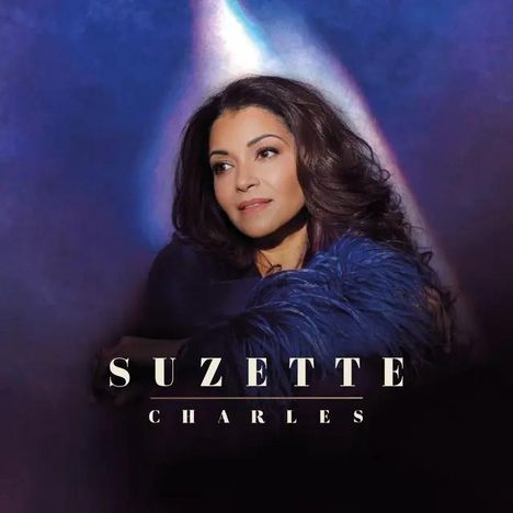 Text: "SUZETTE CHARLES". Frau mit dunklen, gewellten Haaren, in einen blauen Federmantel gehüllt, auf violettem Hintergrund.