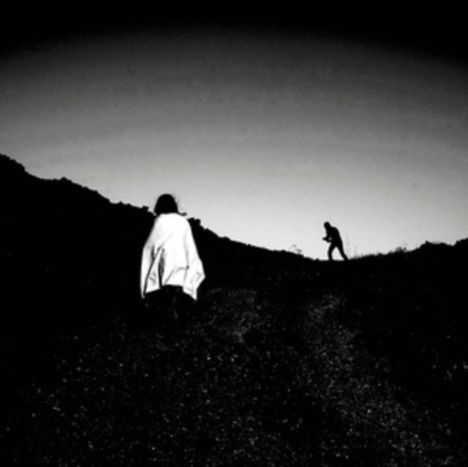 Eine dunkle, bergige Landschaft, vorne eine Person mit Umhang, hinten eine weitere Figur im Gegenlicht.