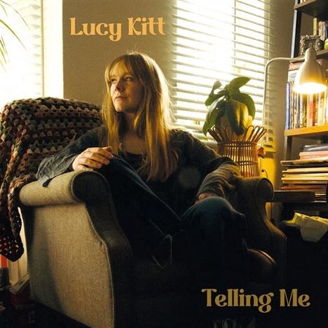 Text: "Lucy Kitt" und "Telling Me". Eine Frau sitzt nachdenklich in einem Sessel, umgeben von Büchern und einer Pflanze.