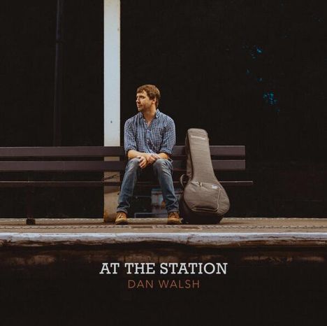 "At the Station" und "Dan Walsh" in Text. Ein Mann sitzt auf einer Bank neben einer Gitarrentasche, umgeben von Dunkelheit.