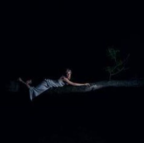 Ein Mensch in einem grauen Kleid liegt waagerecht auf einem breiten Ast im Dunkeln, neben einem kleinen Baum.