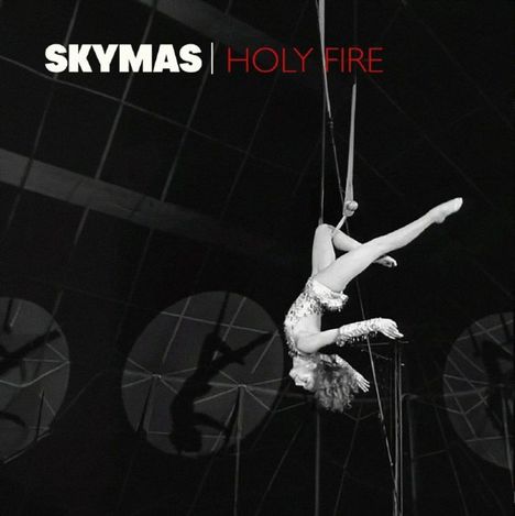 Text: "SKYMAS | HOLY FIRE". Eine Akrobatin in einem schimmernden Outfit schwebt kopfüber an Tüchern in einem Zirkuszelt.