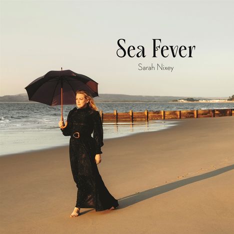 "Sea Fever, Sarah Nixey." Eine Frau in schwarzem Kleid mit Regenschirm spaziert barfuß am Strand entlang.
