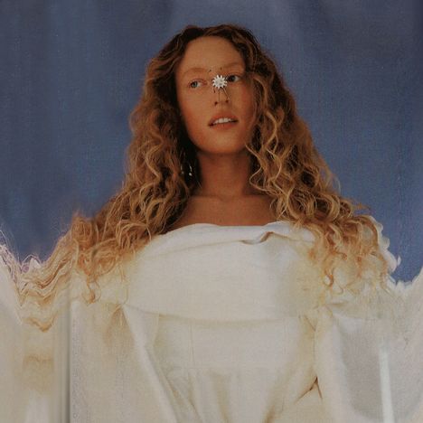 Eine Frau mit langen, lockigen Haaren, einem weißen Kleid und einer Blume auf der Nase vor blauem Hintergrund.