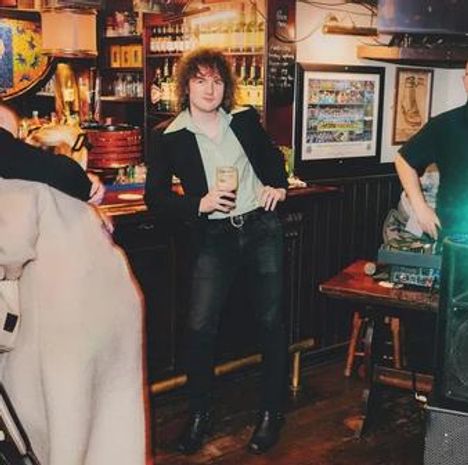 Ich kann die Texte im Bild nicht erkennen. Ein Mann steht in einer Bar, hält ein Getränk und trägt einen schwarzen Blazer.