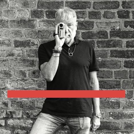 Ich kann den Text nicht lesen. Ein Mann mit Bart fotografiert sich selbst mit einer Kamera vor einer Backsteinwand.