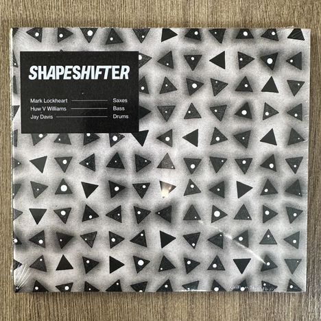 "SHAPESHIFTER" und Bandmitglieder auf Schwarz vor grauem Hintergrund mit schwarzen Dreiecken und weißen Punkten.