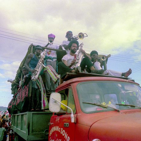 "GERMAN & SONS". Musiker mit Blasinstrumenten auf einem roten Lastwagen vor bewölktem Himmel.