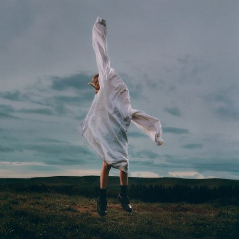 Eine Person in einem weiten, weißen Kleid springt auf einer grünen Wiese bei bewölktem Himmel hoch.
