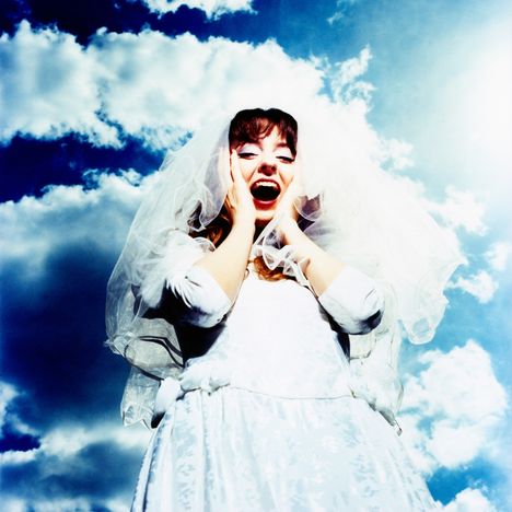 Keine Texte sichtbar. Eine lachende Braut mit Schleier und Kleid, Hände an den Wangen, vor blauem Himmel mit Wolken.