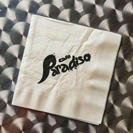Text: "Café Paradiso" in dynamischem Schriftzug auf einer Serviette. Hintergrund mit kreisförmigem Muster.