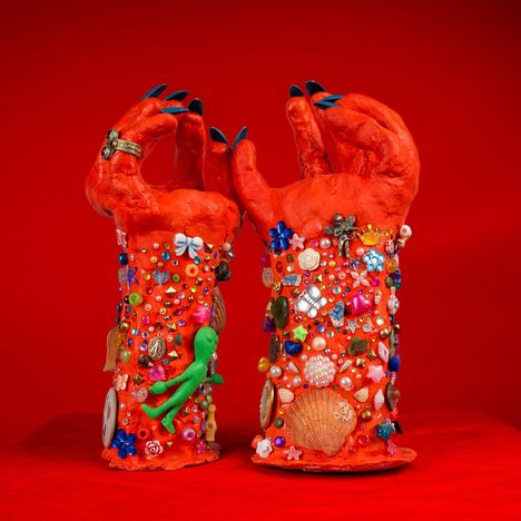 Eine Skulptur von zwei roten Händen, verziert mit bunten Perlen, Muscheln und kleinen Objekten auf rotem Hintergrund.