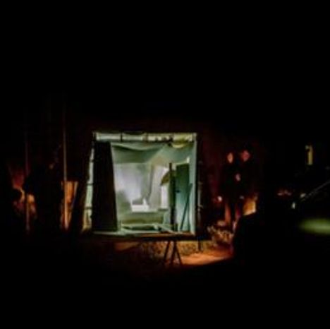 Kein Text erkennbar. Im Fokus steht eine beleuchtete Installation in einem dunklen Raum, umgeben von Personen im Schatten.