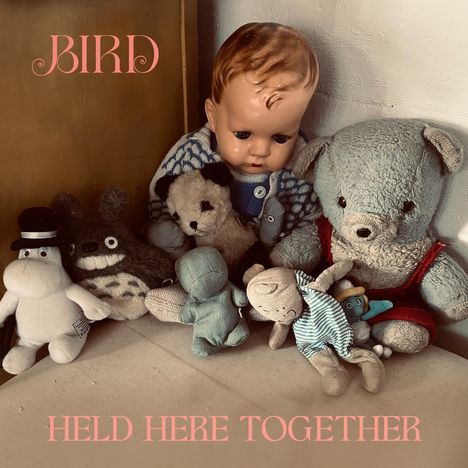 Der Text lautet: "BIRD" und "HELD HERE TOGETHER". Eine Sammlung von Plüschtieren umgibt eine alte Babypuppe.