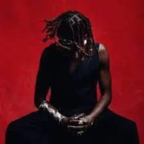 Keine erkennbaren Texte. Person mit Zöpfen sitzt, rote Wand im Hintergrund, Hände gefaltet.