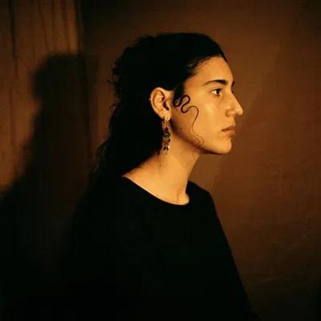 Es gibt keinen Text. Profil einer Person mit dunklen Locken, im Scheinwerferlicht, vor dunklem Hintergrund.