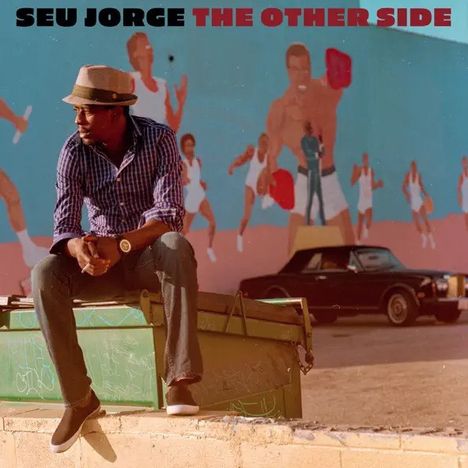 Oben steht "SEU JORGE THE OTHER SIDE". Ein Mann mit Hut sitzt auf einem Container vor einer bemalten Wand.