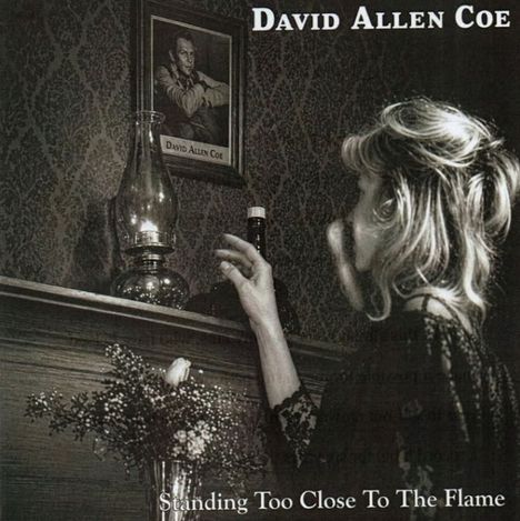 Texte: "David Allen Coe, Standing Too Close To The Flame". Eine Frau zündet ein Streichholz auf einem Kaminsims an.